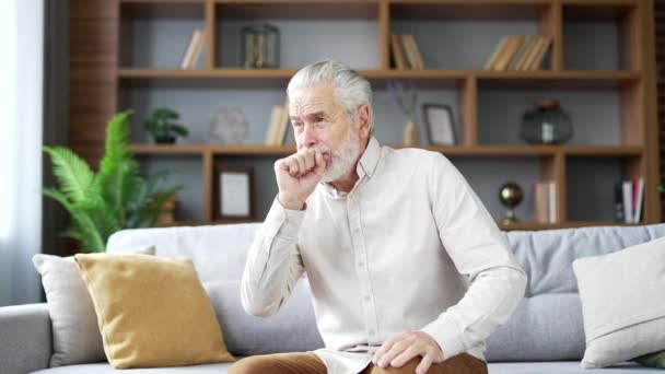 高齢の灰色の毛髪の男は 自宅のリビングルームでソファーに座っている間咳をします 風邪やインフルエンザの病気の成熟した祖父は 彼の胸に手を握ります 彼は高熱または気管支炎を持っています — ストック動画