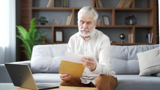 快乐的老年人 白发苍苍的老人 坐在客厅的沙发上 阅读着有好消息的信 快乐的笑着的成年雄性 满足于愉快的通知 庆祝成功 — 图库视频影像