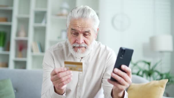 快乐的老年人坐在家里的沙发上 在智能手机上做网上购物 输入信用卡号码 笑着的成年男性会进行购买 并对打折 销售感到满意 — 图库视频影像