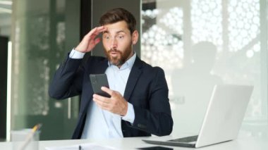 Resmi takım elbiseli mutlu iş adamı iş yerinde otururken akıllı telefondan harika haberler aldı. Neşeli bir girişimci cep telefonundan hoş bir mesaj okur ve başarıyı kutlar.