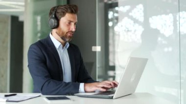 Modern ofisteki çalışma masasında otururken dizüstü bilgisayarda yazan kablosuz kulaklıklarla meşgul bir işadamı. Resmi takım elbiseli yakışıklı girişimci müzik dinlerken bir proje üzerinde çalışıyor