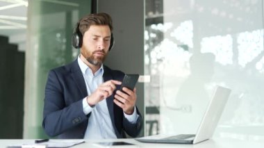 Kulaklıklı yakışıklı iş adamı iş yerinde oturmuş, rahatlatıcı müzik dinliyor. Mutlu girişimci akıllı telefondan çalma listesini açar ve rahatlamanın ve doğanın gürültüsünün tadını çıkarır.