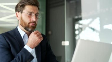 Resmi takım elbiseli ciddi düşünceli bir işadamı iş yerindeki bir laptopta çalışıyor. Gözlüklü sakallı adam bir sorunu çözmeyi düşünüyor, projeyle meşgul. Kapat.