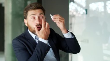 İş yerinde otururken göz yorgunluğu çeken resmi takım elbiseli bir iş adamı. Yorgun bir girişimci, bir göz doktorunun önerdiği görüşü geliştirmek için göz ilacı enjekte eder. Kapat.