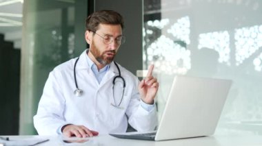 Hastane kliniğinde laptopla konuşan kendine güvenen bir doktor. Erkek tıp doktoru ofisteki bir konferansta uzaktan iletişim kurar ya da bir hastayla çevrimiçi istişarede bulunur.