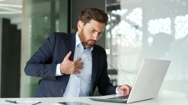 正式なスーツのビジネスマンは ビジネスオフィスの職場に座っているラップトップに取り組んでいる間 心臓発作を起こします 病気の男性は胸の緊張に苦しみ 彼の手で痛みのスポットを保持しています — ストック動画