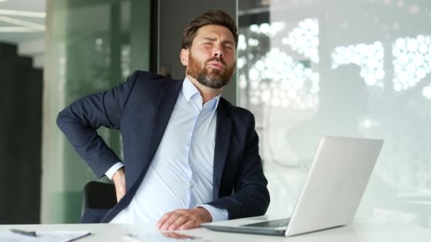 ビジネスオフィスの職場で座っているラップトップに取り組んでいる間 背中の痛みに苦しむ正式なスーツの疲れたビジネスマン Upsetは痛みを伴う腰の筋肉をマッサージします 彼は疲れていて過労しています — ストック動画