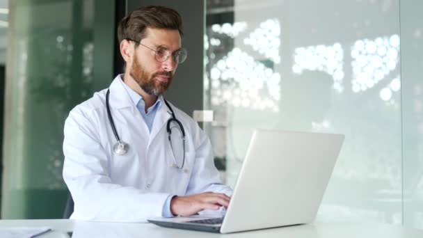 在医院诊所工作时 穿着白色外套的有信心的医生坐在笔记本电脑上工作 医务工作者的医生在网上与病人聊天 写信息或使用计算机应用程序 — 图库视频影像