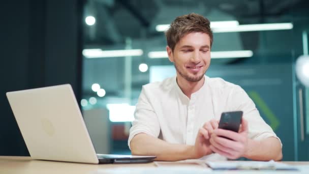 従業員はテキストメッセージでオフィスでチャットしています 彼の友人とソーシャルメディアのウェブをサーフィンする携帯電話のスマートフォンを使用して オンラインで座ってブラウジングする若いハンサムな男性 — ストック動画