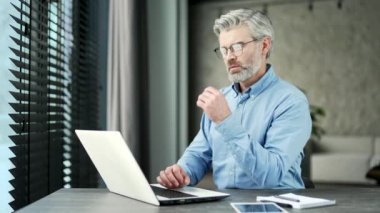 Düşünceli, olgun, gri sakallı iş adamı iş yerindeki bir laptopta çalışıyor. Üst düzey sahibi masajla sohbet yazılımı, bilgisayar uygulamasında bankacılık, yazılım uygulaması kullanarak
