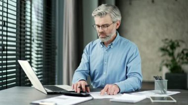 Yetişkin, gri sakallı iş adamı hesap makinesi kullanarak finansal hesaplamalar yapıyor, iş yerinde dizüstü bilgisayarda yazıyor. Kendine güvenen yatırımcı, iş yerinde belgeleri dolduruyor.