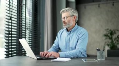 Aşırı çalışılmış gri sakallı iş adamı göz yorgunluğu ve baş ağrısından muzdarip iş yerinde oturan bir dizüstü bilgisayarla çalışıyor. Yorgun adam gözlüğünü ve masajını çıkarıyor, burun köprüsünü ovuyor.