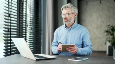 Olgun, gri saçlı iş adamı online video konferansı seyrediyor. Ofiste oturan dizüstü bilgisayara bakarak notlar alıyor. Uzaktan kumandayı dinleyen bir erkek özel öğretmenle konuşuyor.