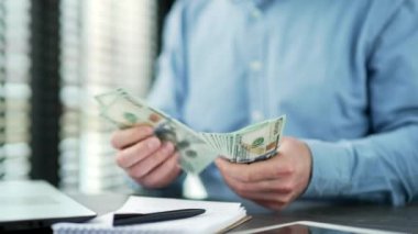 Kapatın. Erkek eller 100 dolarlık banknotları sayar. İş yerindeki masalarda banknotları sayan mavi gömlekli olgun bir iş adamı. Adam parayı elinde tutuyor. Mali başarı kavramı