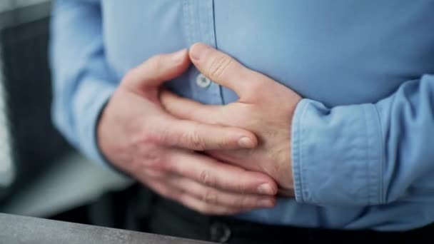 クローズアップ 大量の胃を抱えている男性の手 青いシャツのビジネスマンは ビジネスオフィスの職場に座っている胃の痛みを感じます 病気の男は 血まみれ 胃炎中毒またはスパムに苦しんでいる — ストック動画