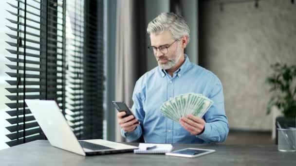 一个头发灰白的老实人 手里拿着一大把钞票和智能手机 坐在办公室里看着摄像机 男人对用手机申请赚钱感到满意 — 图库视频影像
