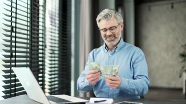 İş yerindeki banknotları sayarak tatmin olmuş gri sakallı iş adamı. Gülümseyen kıdemli girişimci elinde iyi kazançlarla mutlu bir şekilde nakit para tutuyor.