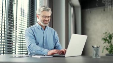 Olgun gri sakallı iş adamı iş yerinde çalışırken dizüstü bilgisayarda çalışıyor. Kıdemli girişimci yazıcı masajla sohbet ediyor bilgisayar uygulamasında bankacılık yapıyor, yazılım uygulaması kullanıyor