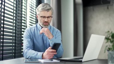 Düşünceli, gri sakallı iş adamı iş yerindeki akıllı telefonuna göz atıyor. Ciddi bir girişimci masaj okur, sohbet eder, uygulama bankacılığı yapar, yazılım uygulaması kullanır