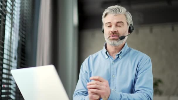 成熟した灰色のひげ付きビジネスマンは オフィスに座っているラップトップを使用してビデオ通話で話しているヘッドセットで 起業家にはビジネスミーティングがあります 上司がオンラインカンファレンスでリモートで話す — ストック動画