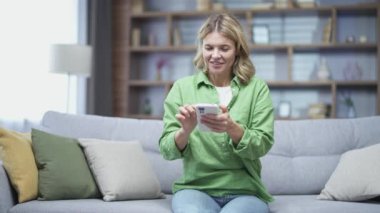 Mutlu olgun bir kadın evdeki koltukta otururken akıllı telefon kullanıyor. Kadın sosyal medyada geziniyor, çevrimiçi mağazada alışveriş yapıyor, sipariş veriyor ya da online bankacılık için ödeme yapıyor