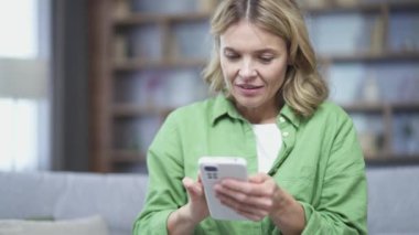 Mutlu olgun bir kadın evdeki koltukta otururken akıllı telefon kullanıyor. Kadın sosyal medyada geziniyor, çevrimiçi mağazada alışveriş yapıyor, sipariş veriyor ya da online bankacılık için ödeme yapıyor