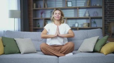 Gözleri kapalı meditasyon yapan olgun bir kadın evdeki oturma odasındaki kanepede nilüfer pozisyonunda oturuyor. Sakin bir kadın günlük giysiler içinde dinleniyor işten sonra yoga egzersizleri yapıyor.