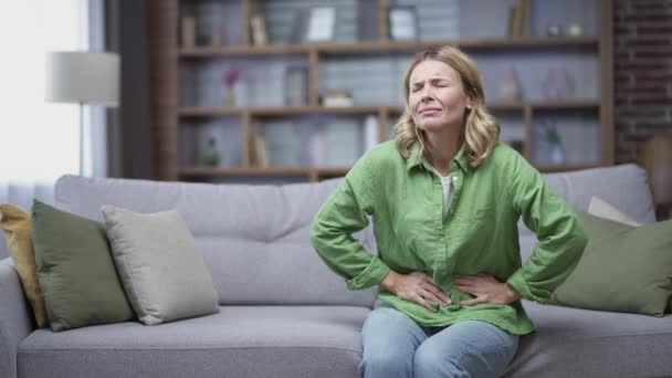 病気の成熟した女性は 自宅のリビングルームでソファーに座っている間に胃痛を感じます 疲れた中年女性急性胃炎または月経痛を感じるスパム中毒や下痢 — ストック動画