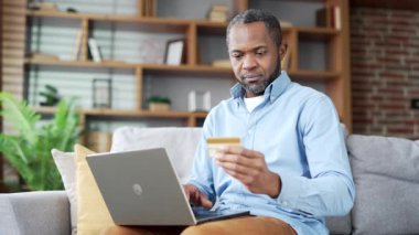 Mutlu Afro-Amerikalı adam online alışveriş yapıyor dizüstü bilgisayarında kredi kartı numarasını yazıyor evdeki kanepede oturuyor. Gülümseyen yaşlı sakallı erkek satın alıyor, terfi ve indirimlerle tatmin oluyor.