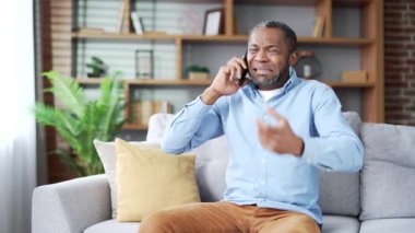 Kızgın, heyecanlı, olgun, Afro-Amerikan erkek akıllı telefondan tartışıyor, evdeki oturma odasında kanepede oturuyor. Sinirli yaşlı zenci adam telefonda çözülmesi gereken sorunlardan yakınıyor.