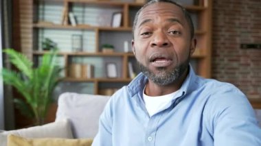 POV Olgun Afro-Amerikalı bir adam video konuşmasında akıllı bir telefonla konuşuyor. Evdeki kanepede oturan kameraya bakıyor. Gülümseyen son sınıf sakallı bir erkek arkadaşı ya da iş arkadaşıyla internette sohbet eder.