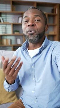 POV dikey video. Olgun Afro-Amerikalı bir adam video konuşmasında akıllı telefon kullanarak evde oturan kameraya bakıyor. Gülümseyen kıdemli işadamı iş ortağıyla internette sohbet ediyor.