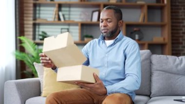 Heyecanlı Afro-Amerikan erkek müşteri paketini evde açıyor. Mutlu son sınıf sakallı erkek alıcı koltukta oturuyor. E-ticaret Ekspres dağıtım hizmetleri konseptinin tatmin olmuş müşterisi