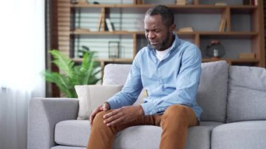 Olgun Afro-Amerikan erkeği evdeki oturma odasında kanepede otururken şiddetli diz eklem ağrısı çekiyor. Yaşlı erkek masajı, bacak kasları, yürüyemiyor, hastalık, kramp, romatizma.
