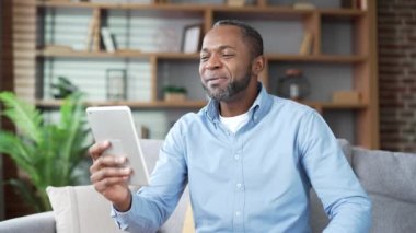 Olgun Afro-Amerikalı bir adam, evdeki oturma odasındaki kanepede oturup tablet ekrana bakarken bir selfie çekiyor. Gülümseyen yaşlı sakallı erkek bir arkadaşıyla internette sohbet ediyor.