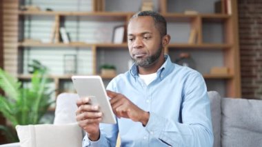 Olgun Afro-Amerikan sakallı adam evdeki oturma odasında kanepede otururken dijital tablet kullanıyor. Erkek sosyal ağlara takılır, çevrimiçi alışveriş yapar, müşteriyle sohbet eder, mesaj yazar veya okur