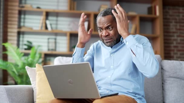 繊細な成熟したアフリカ系アメリカ人のひげをした男は 自宅のリビングルームでソファーに座っている間 ノートパソコンで悪いニュースを読みます 心配したうつ病の男性は コンピュータ上の否定的なメッセージをレビューしながら悲しいです — ストック動画