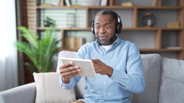 兴奋的成熟的非洲裔美国男性在无线耳机中坐在客厅的沙发上观看田径比赛 快乐的老年人在感情上为最喜欢的球队欢呼 — 图库视频影像