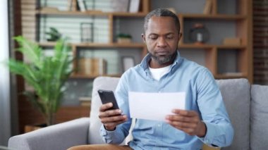 Olgun Afro-Amerikalı adam evde otururken akıllı telefon kullanarak faturaları harcamak için mobil uygulama kullanıyor. Erkek, ev ekonomisini hesaplar ve verileri sohbet robotuna aktarır. Ödemesi kolay ve basit.