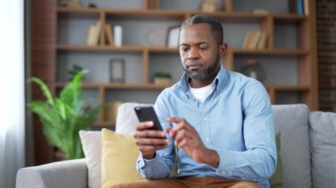 Olgun Afro-Amerikan sakallı adam evdeki oturma odasındaki koltukta akıllı telefon kullanıyor. Üst düzey erkek sosyal ağlara takılır, çevrimiçi bir mağazadan alışveriş yapar, sohbet eder, yazar veya mesajları okur