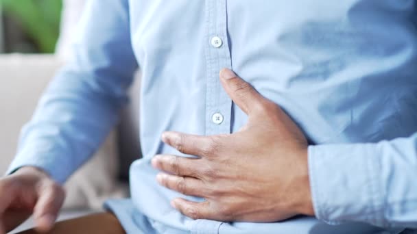 クローズアップ 大量の胃を抱えている男性の手 青いシャツの成熟したアフリカ系アメリカ人男性は 自宅のリビングルームでソファーに座っている胃の痛みを感じます 病気の人は 胃炎またはスパムに苦しんでいます — ストック動画