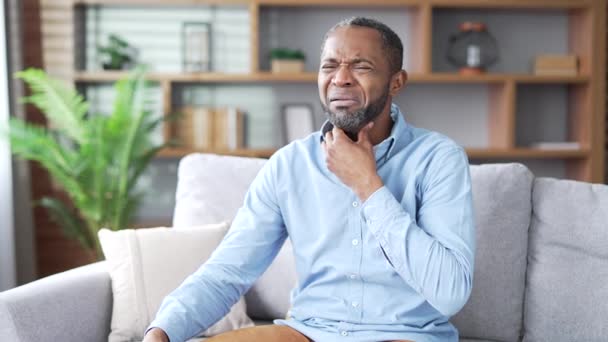 病気の成熟したアフリカ系アメリカ人男性は 自宅のリビングルームでソファーに座っている間に喉の痛みに苦しんでいます 上のひげ付き男性は風邪 ウイルス インフルエンザを持っています 痛みの場所をマッサージするUpsetブラックマネージャー — ストック動画