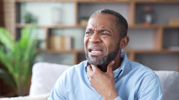 病気の成熟したアフリカ系アメリカ人男性は 自宅のリビングルームでソファーに座っている間に喉の痛みに苦しんでいます 高齢男性は風邪 ウイルス インフルエンザを患っています 痛みの場所をマッサージするウプセットブラックマン クローズアップ — ストック動画