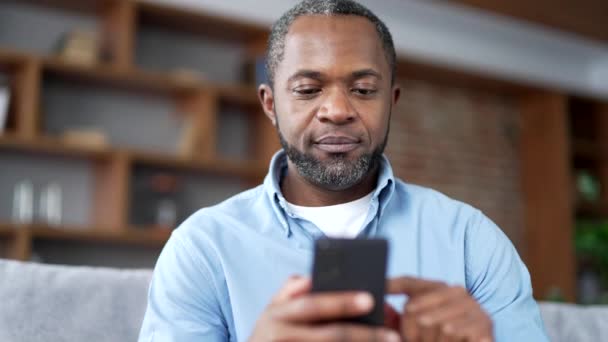 成熟的非洲裔美国胡子男人坐在客厅的沙发上 用智能手机 年长的男性浏览社交网络 在网上商店购物 写信息 靠近点 — 图库视频影像