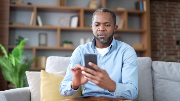自宅のリビングルームにソファーに座っているスマートフォンでソフトウェアやサービスに問題を抱えている成熟したアフリカ系アメリカ人男性 心配した失望した男性が悪いインターネット接続について不満を言います — ストック動画