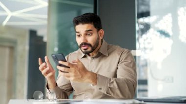 Sinirli iş adamı, iş yerindeki bir masada otururken akıllı telefondan kötü haberler okur. Endişeli erkek işçi cep telefonundaki olumsuz bilgileri incelerken üzgün