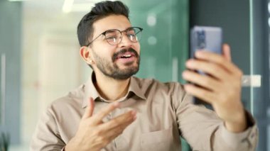 Gözlüklü, gülümseyen sakallı bir iş adamı modern bir ofisteki akıllı telefon ekranına bakarak selfie çekiyor. Yakışıklı bir işçi müşterisiyle veya iş ortağıyla çevrimiçi sohbet eder.