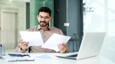Gözlüklü gülümseyen finansör, iş yerinde otururken incelediği mali raporun sonuçlarından memnun. Girişimci işadamı olumlu göstergelerden memnun