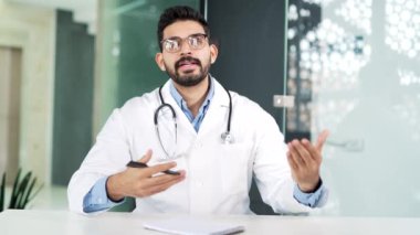 Webcam görüntüsü. Doktor, hastane kliniğindeki ofiste kameraya bakarken görüntülü konuşuyor. Güvenilir erkek doktor konferansta uzaktan iletişim kuruyor ya da online danışmanlık yapıyor.
