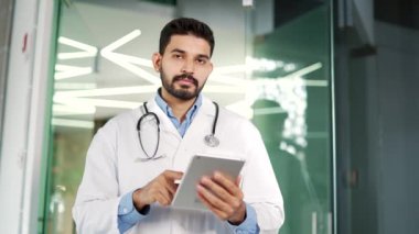 Steteskoplu beyaz önlüklü gülümseyen sakallı tıp doktorunun portresi hastane kliniğinde elinde tabletle kameraya bakıyor. Pozitif yakışıklı doktor vesikalığı.